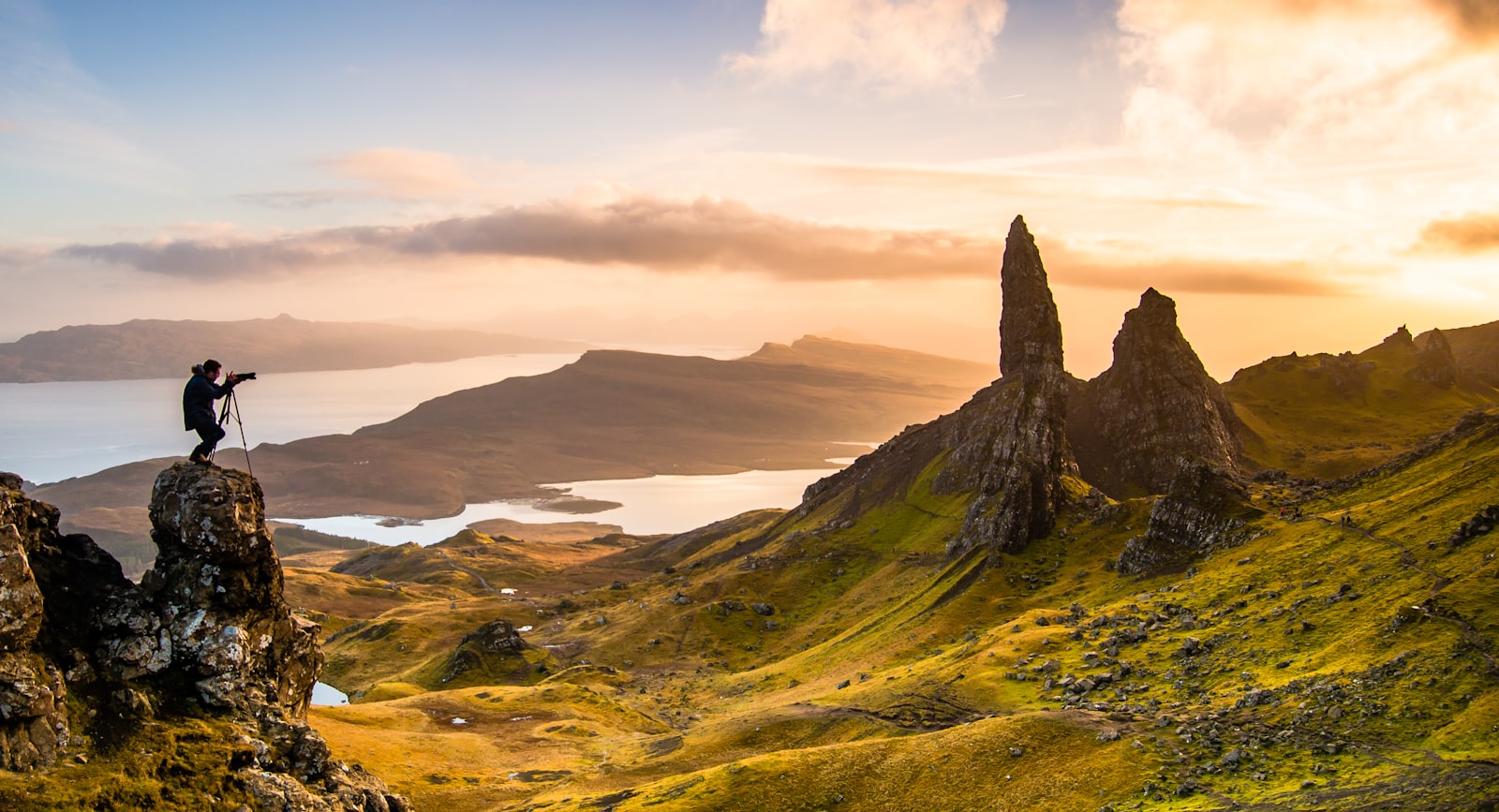 A photographer on a rock outcrop facing the pinnacles of the Old Man of Storr with lochs and the sound below